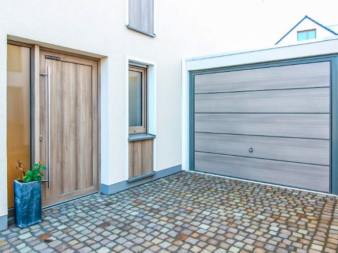 Hauseingang eines Neubaus mit Haustür in Holzoptik, Garagentor und gepflasterter Zufahrt.
